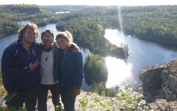 Three people pose for a photo with their arms around each other. They appear to be standing on a lookout, over a bending body of water and green woods.